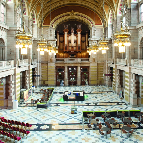 An internal view of the Kelvingrove Main Hall taken from a first storey balcony shows a cavernous, vaulted, 3 storey space adorned with carved sandstone walls, a patterned polished-stone floor and a dozen glass and golden chandaliers. A carved stone banister is visible around the entire first-storey balcony.  A huge pipe organ is visible on the balcony at the far, opposite end of the room, large wooden doors are visible beneath it. Small tables and seats are gathered together in parts of the large floor.
