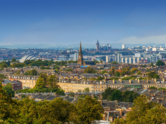 Sunny cityscape with the Gothic revival-style spire of the University of Glasgow's main building, surrounded by greenery and buildings of the West End, including the modern buildings of the SEC Armadillo and the OVO Hydro arena